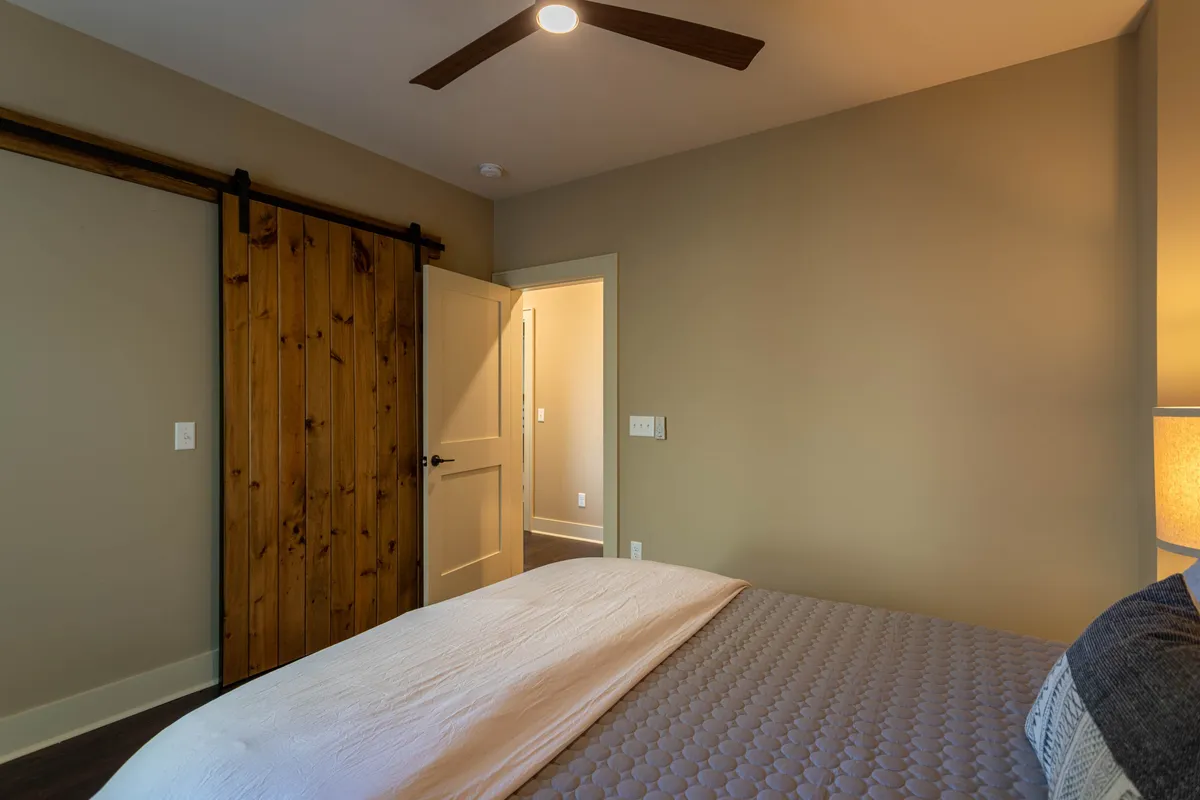This interior image showcases a cozy bed accented by a wooden sliding door and a bedside lamp. The soft natural light filtering in creates a tranquil atmosphere, inviting relaxation. The space looks spacious and thoughtfully designed, suggesting a suite or main bedroom retreat.