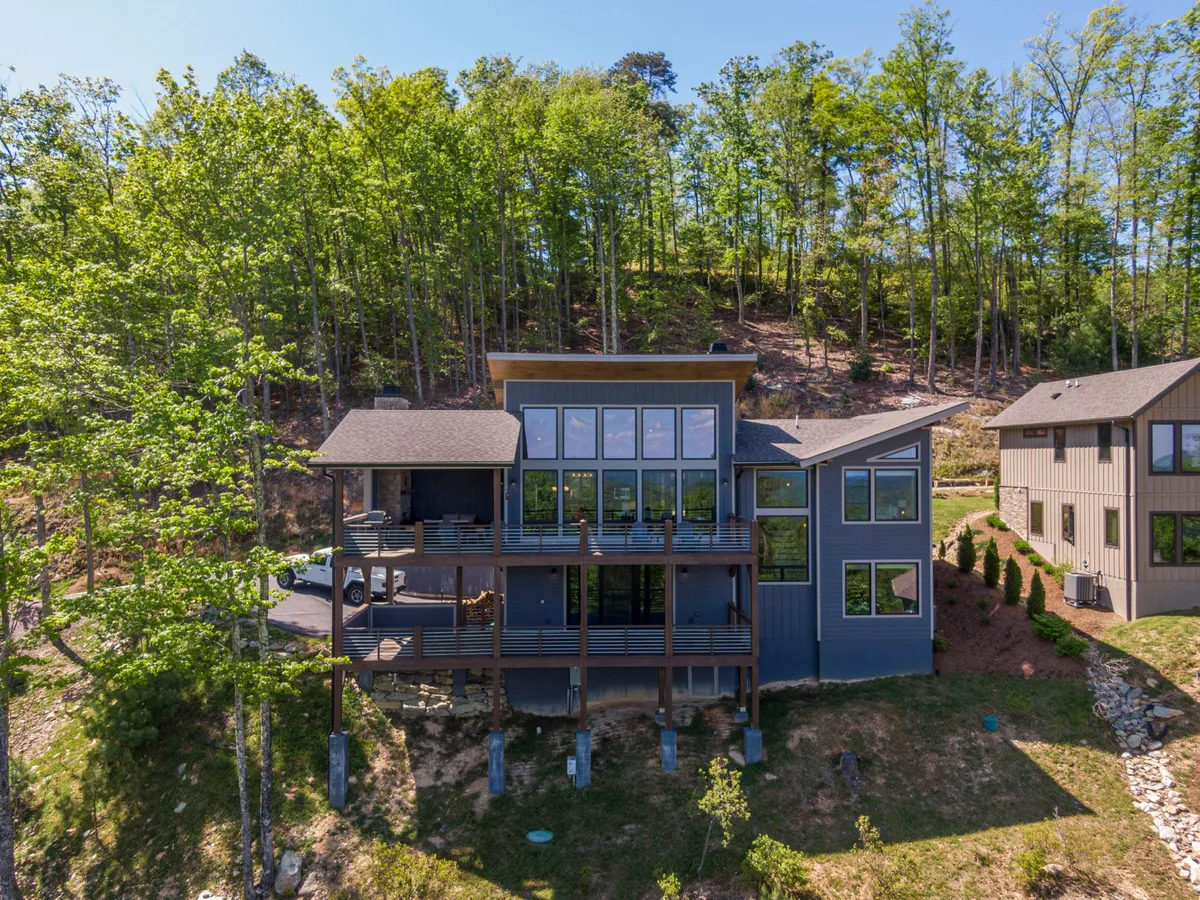 This striking exterior shot showcases a modern mountain home with expansive glass windows, a spacious deck, and lush forest surroundings. The contemporary design is complemented by panoramic sunset views, inviting outdoor lounging, and a seamless connection to nature.