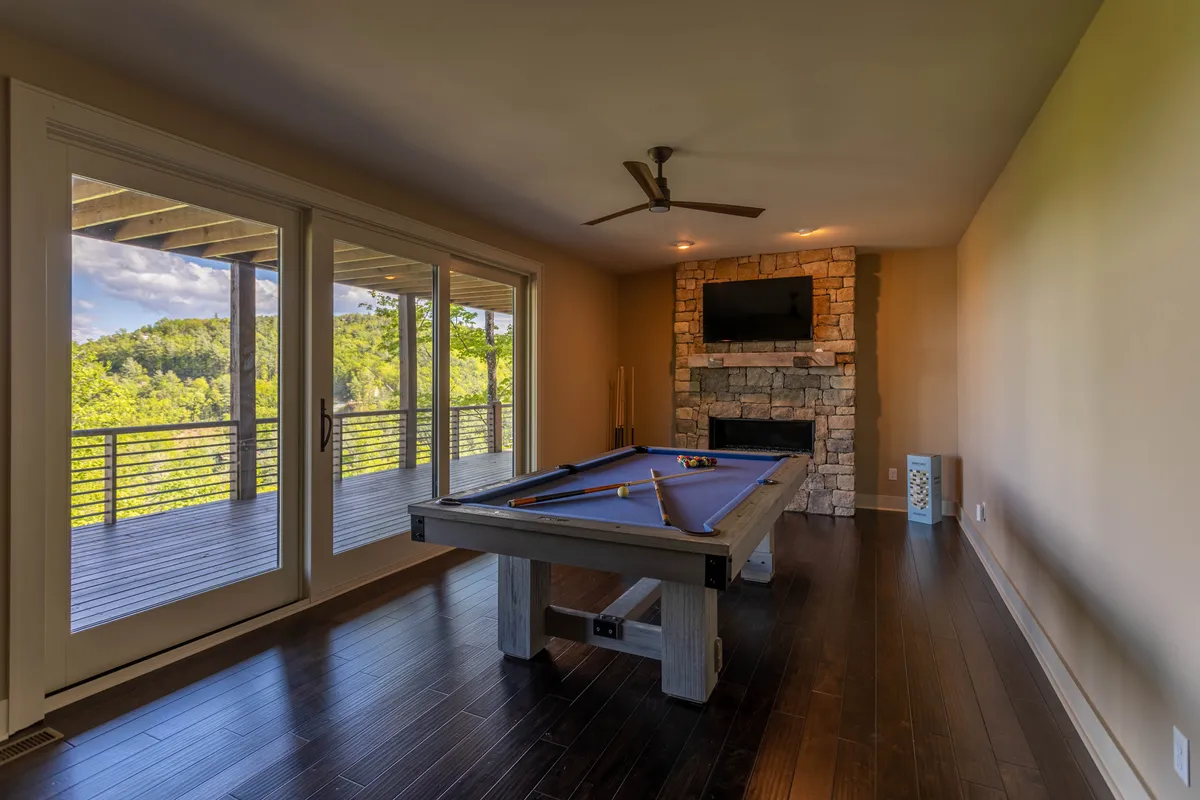A vibrant game room with a sleek pool table as its centerpiece, set against a backdrop of a stone fireplace and expansive windows that let in natural light. The ambience is playful and relaxed, with space for socializing and unwinding after a day of activities.