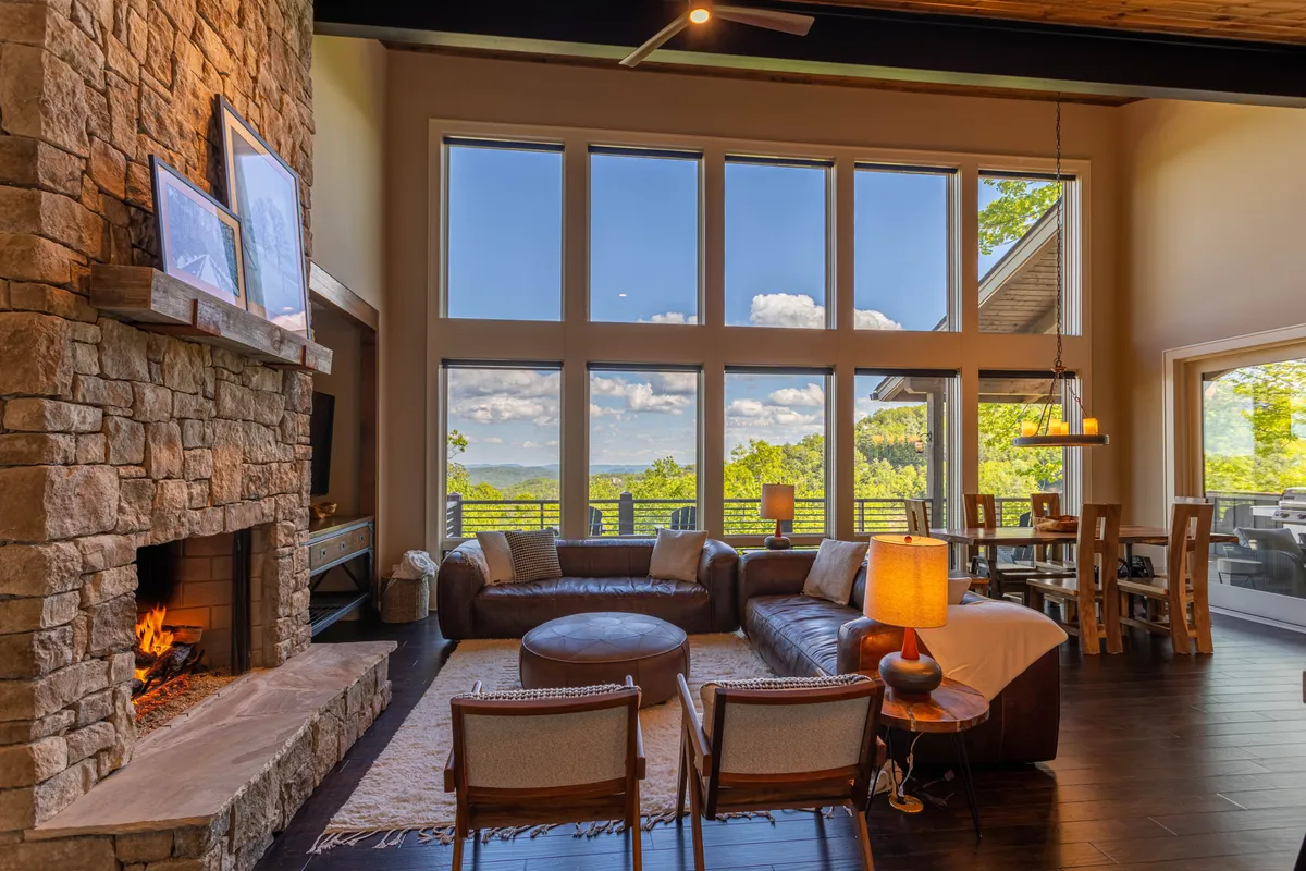 This image features a stunning open-concept living area with a stone fireplace, large panoramic windows that frame mountain views, a plush leather sofa, and a wooden dining table. The natural light fills the space, highlighting the cozy yet grand atmosphere.