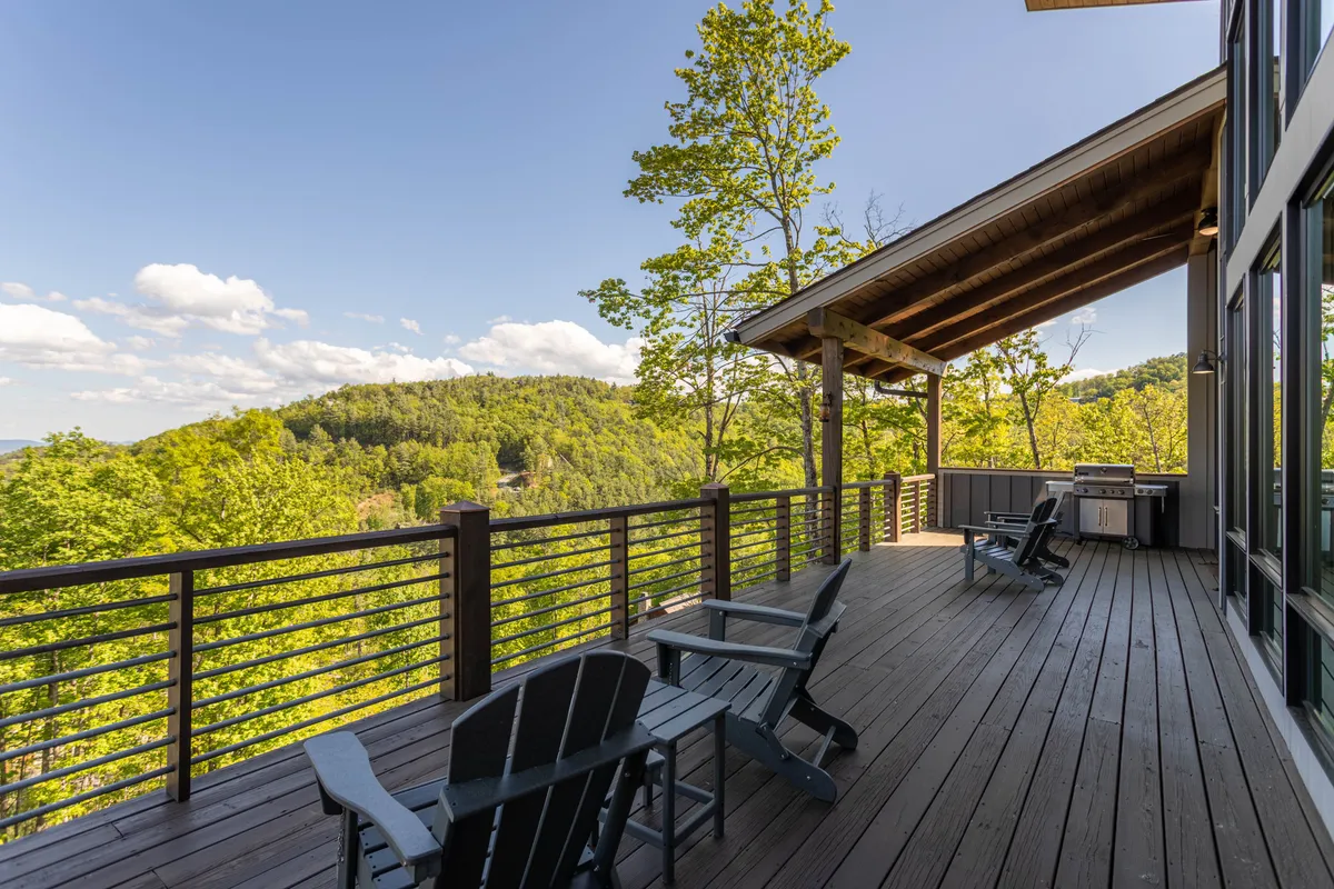 A covered deck with plush wicker seating overlooks rolling hills, featuring a barbecue area and plenty of space to gather. The setting highlights the seamless connection between indoor and outdoor living, with sunrise and stargazing opportunities.