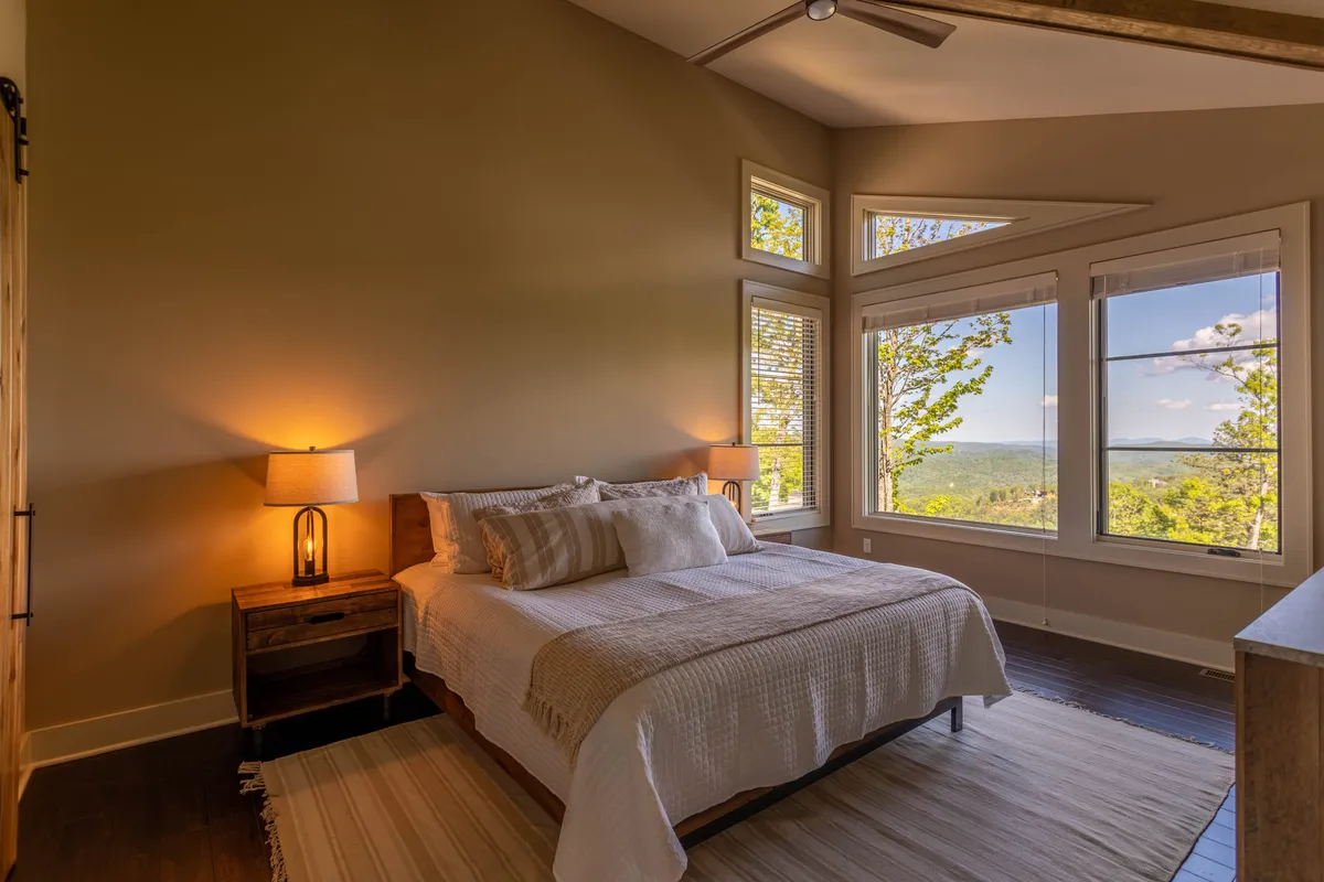 A tranquil bedroom scene with a comfortable bed positioned near large windows. Soft lighting, a bedside lamp, and serene views through the window contribute to a restful, inviting environment ideal for relaxation or enjoying the landscape.