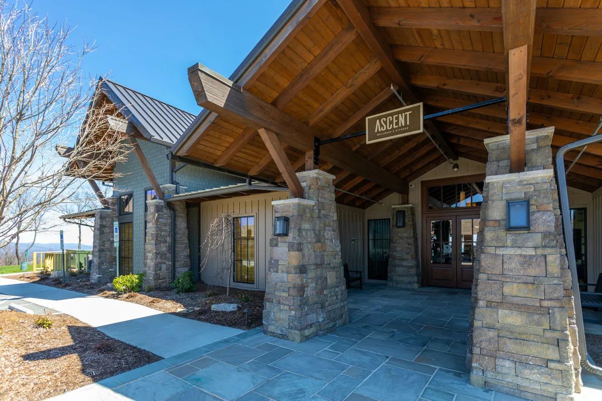 The photo highlights the inviting entrance to the Ascent Wellness & Fitness Center, identifiable by the 'ASCENT' sign. It features striking wooden beams and stone pillars, with a spacious outdoor area for gathering, all set against a serene backdrop.