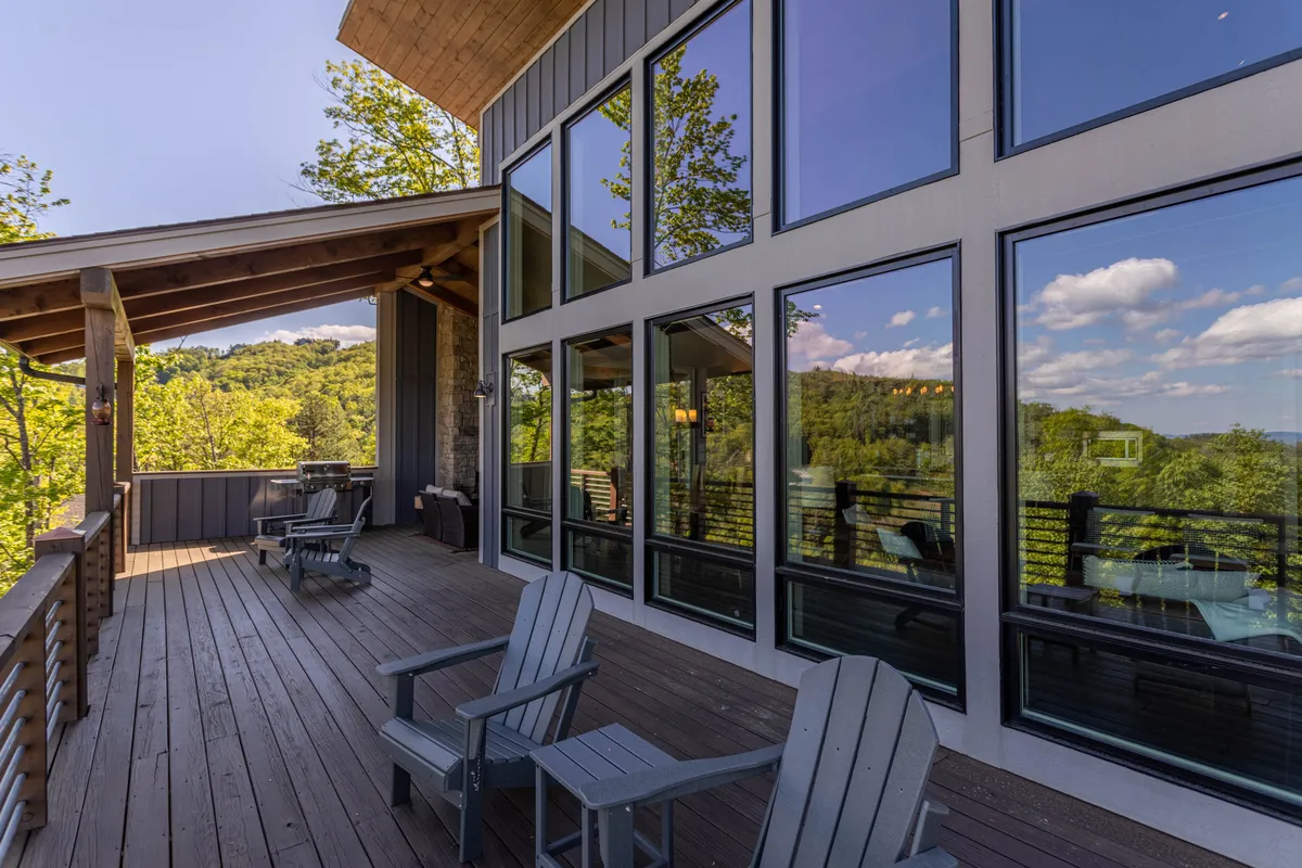 This exterior shot presents a spacious wooden deck with Adirondack chairs, large glass windows reflecting the greenery, and panoramic nature views. The scene is tranquil, perfect for morning coffee or evening relaxation while enjoying the mountain vistas.