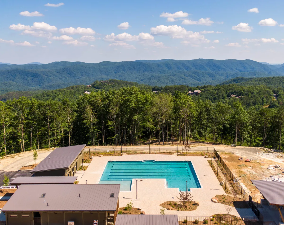 A picturesque outdoor swimming pool with sweeping mountain views and lush trees surrounding the area. People can be seen swimming, sunbathing, and gathering for conversation as the sun sets, creating a relaxed yet luxurious setting.