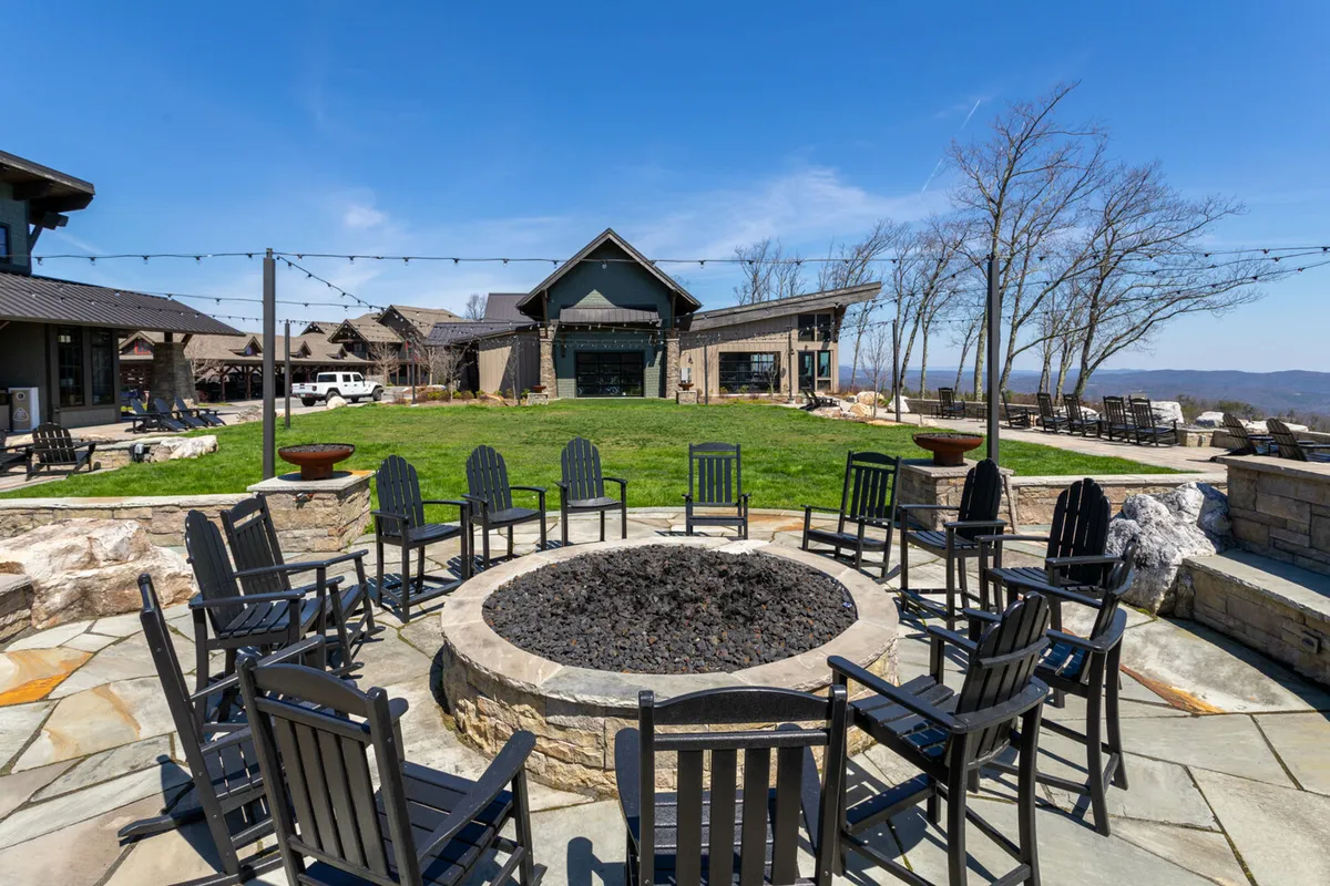 This vibrant exterior scene features a cozy fire pit surrounded by comfortable chairs, with the main building serving as an inviting backdrop. String lights overhead set the mood for social gatherings, while the sunset sky enhances the warm, welcoming atmosphere. People are seen enjoying each other's company, suggesting a perfect setting for group activities and family fun.